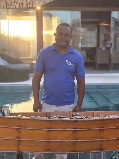 Atendente de camisa Point da Ostra atrás de um barco de madeira cheio de ostras no gelo, em atendimento de evento ao entardecer.