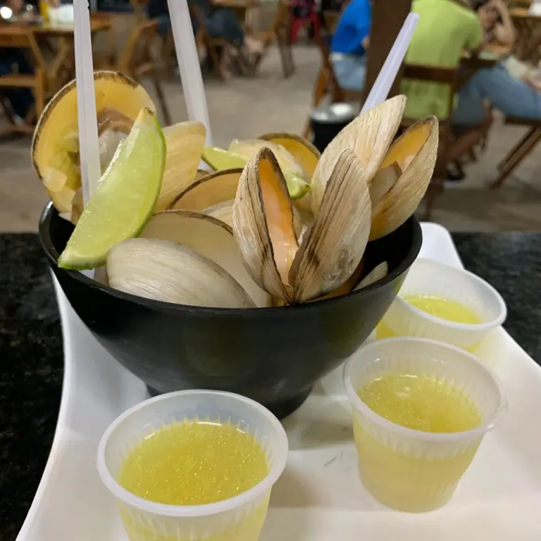 Porção de lambretas abertas na concha, com rodelas de cebola e um gomo de limão, servida em tigela preta, com potinhos de molho ao lado.