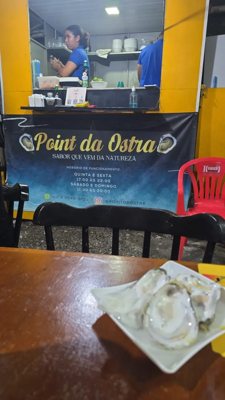O ponto do Point da Ostra à noite, no food park: banner com o nome, a tagline e os horários de funcionamento na frente do balcão amarelo, equipe de camisa azul atendendo, e um prato de ostras na mesa em primeiro plano.